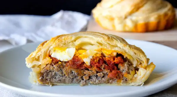 Porción de pastel de carne jugoso con huevo duro y salsa de tomate, envuelto en masa dorada.