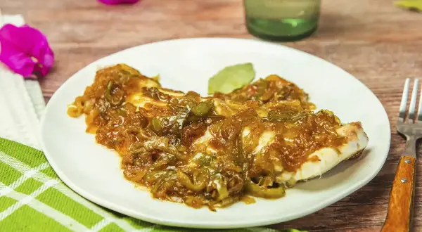 Un plato de urta a la roteña, con pescado blanco cubierto de cebolla y pimientos pochados, servido en una mesa de madera.
