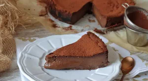 Una rebanada de tarta de chocolate, cubierta de cacao en polvo, reposa en un plato blanco. El postre parece cremoso y delicioso.