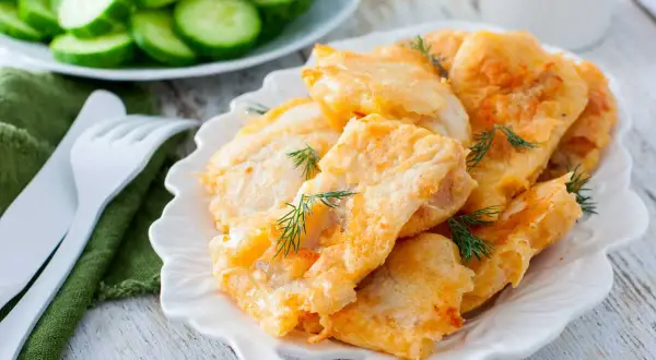 Delicioso cachopo de merluza rebozado, servido en plato blanco con eneldo fresco. Acompañado de rodajas de pepino.