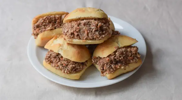 Cinco montaditos de pringa, rellenos de carne desmenuzada y especias, apilados en un plato blanco sobre un mantel de lino.