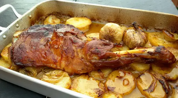 Paletilla de cordero lechal al horno estilo segoviano, dorada y jugosa, acompañada de patatas y ajos asados en su jugo.