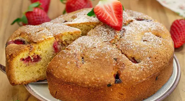 Delicioso bizcocho de fresa, espolvoreado con azúcar glas y decorado con una fresa fresca. Se ve una porción cortada, revelando trozos de fresa en su interior.