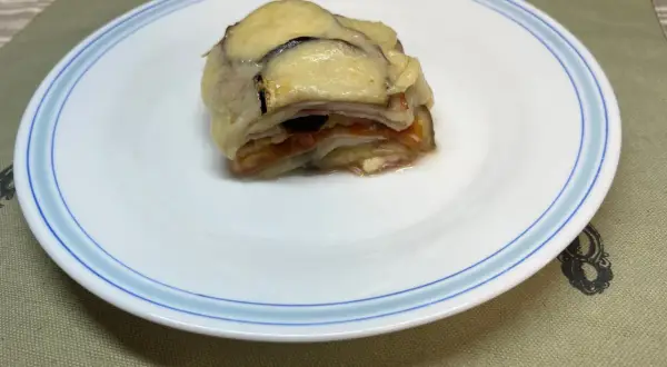 Un delicioso pastel de berenjenas gratinado con queso, servido en un plato blanco con borde azul.