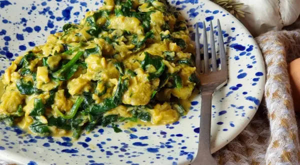 Un delicioso revuelto de espinacas cremoso, servido en un plato con salpicaduras azules. Un tenedor plateado descansa a un lado, listo para disfrutar.