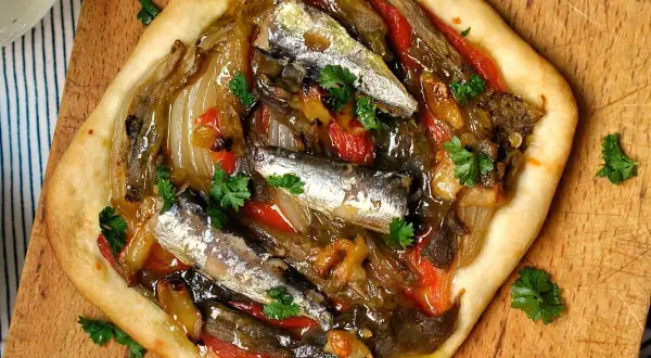 Deliciosa coca de verduras con sardinas y perejil fresco, servida sobre tabla de madera.