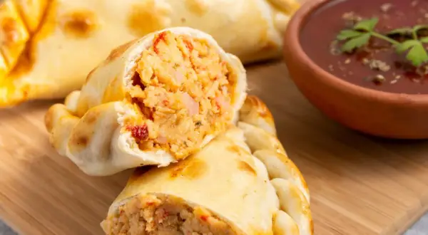 Empanada de pollo recién horneada, cortada por la mitad, revela un relleno jugoso. A su lado, un cuenco de salsa roja con perejil.