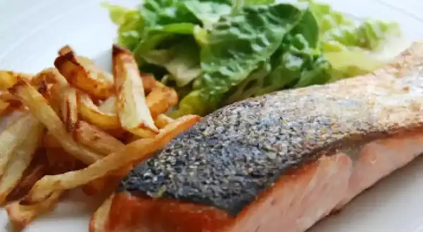 Un delicioso salmón a la plancha con piel crujiente, acompañado de patatas fritas doradas y una fresca ensalada verde.