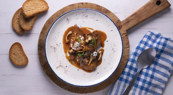 Fricandó de vedella amb bolets i una salsa espessa, servit en un plat blanc amb vores blaves, acompanyat de torradetes.