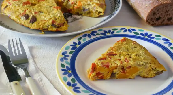 Deliciosa tortilla de berenjena con pimientos y calabaza, servida en un plato decorado. Acompañada de pan rústico y cubiertos.