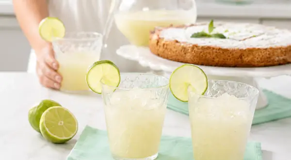 Refrescante granizado de limón servido en vasos con rodajas de lima, junto a limones frescos y un pastel.