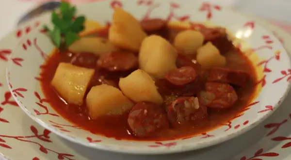 Un plato humeante de patatas a la riojana, con trozos de chorizo y patata en una salsa rojiza, decorado con una ramita de perejil.