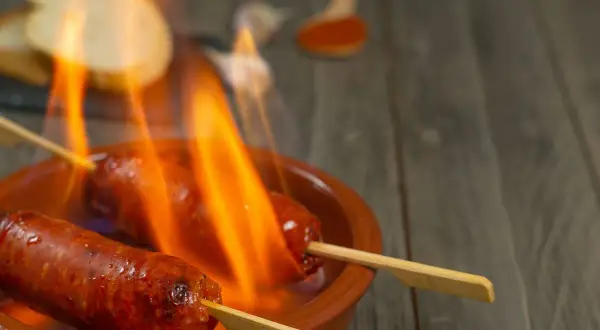 Chorizo al infierno, dos salchichas en brochetas ardiendo en una cazuela de barro, con pan y pimentón al fondo.
