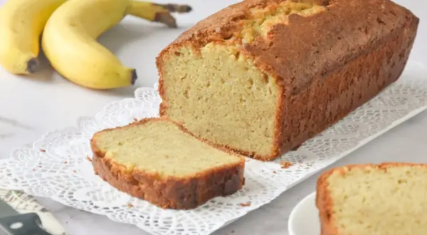 Un delicioso bizcocho de plátano recién horneado, con una rebanada cortada y dos plátanos amarillos al fondo.