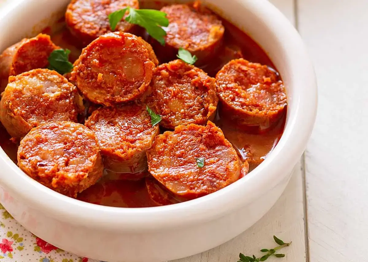 Chorizos a la sidra jugosos y dorados en una salsa rojiza, servidos en un cuenco blanco.