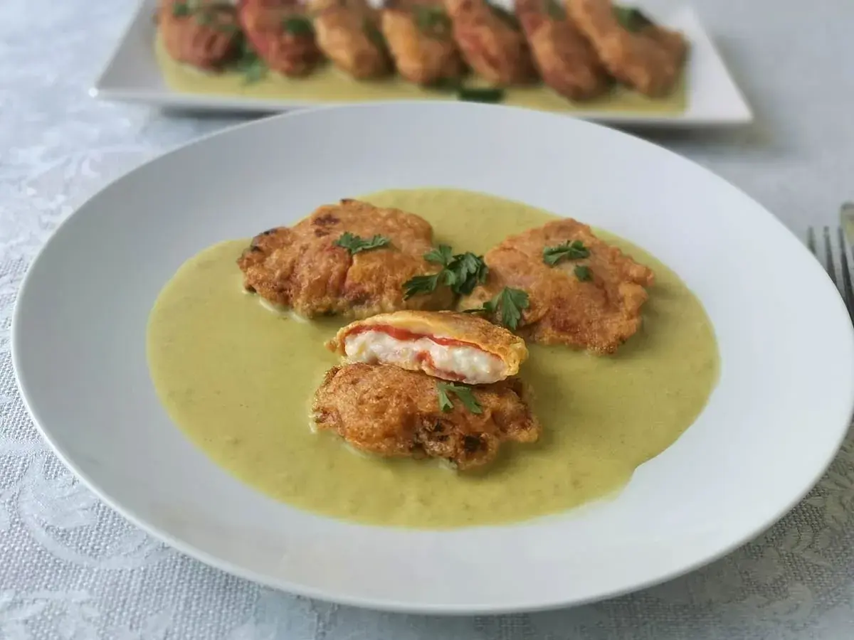 Pimientos piquillo rellenos rebozados, servidos en una cremosa salsa verde. Uno está cortado, mostrando su interior.