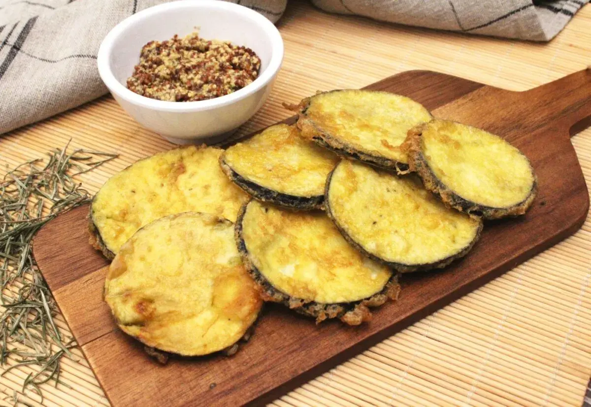 Berenjenas rebozadas con harina y huevo, doradas y crujientes, servidas en tabla de madera con un cuenco de mostaza.
