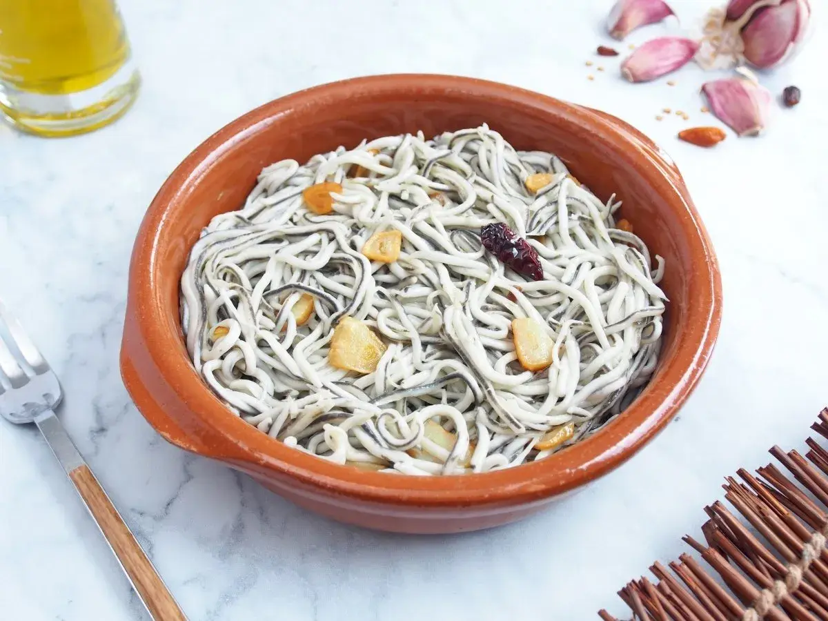 Un cuenco de barro con gulas al ajillo, salteadas con ajo y guindilla. Se ve un tenedor y una botella de aceite de oliva.