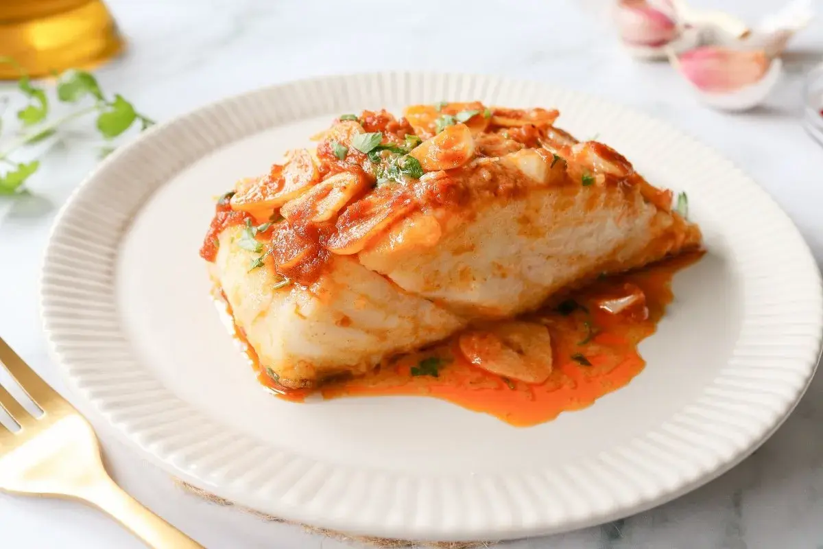 Bacalao a la llauna jugoso, cubierto de salsa de tomate y ajo laminado, servido en plato blanco con tenedor dorado.