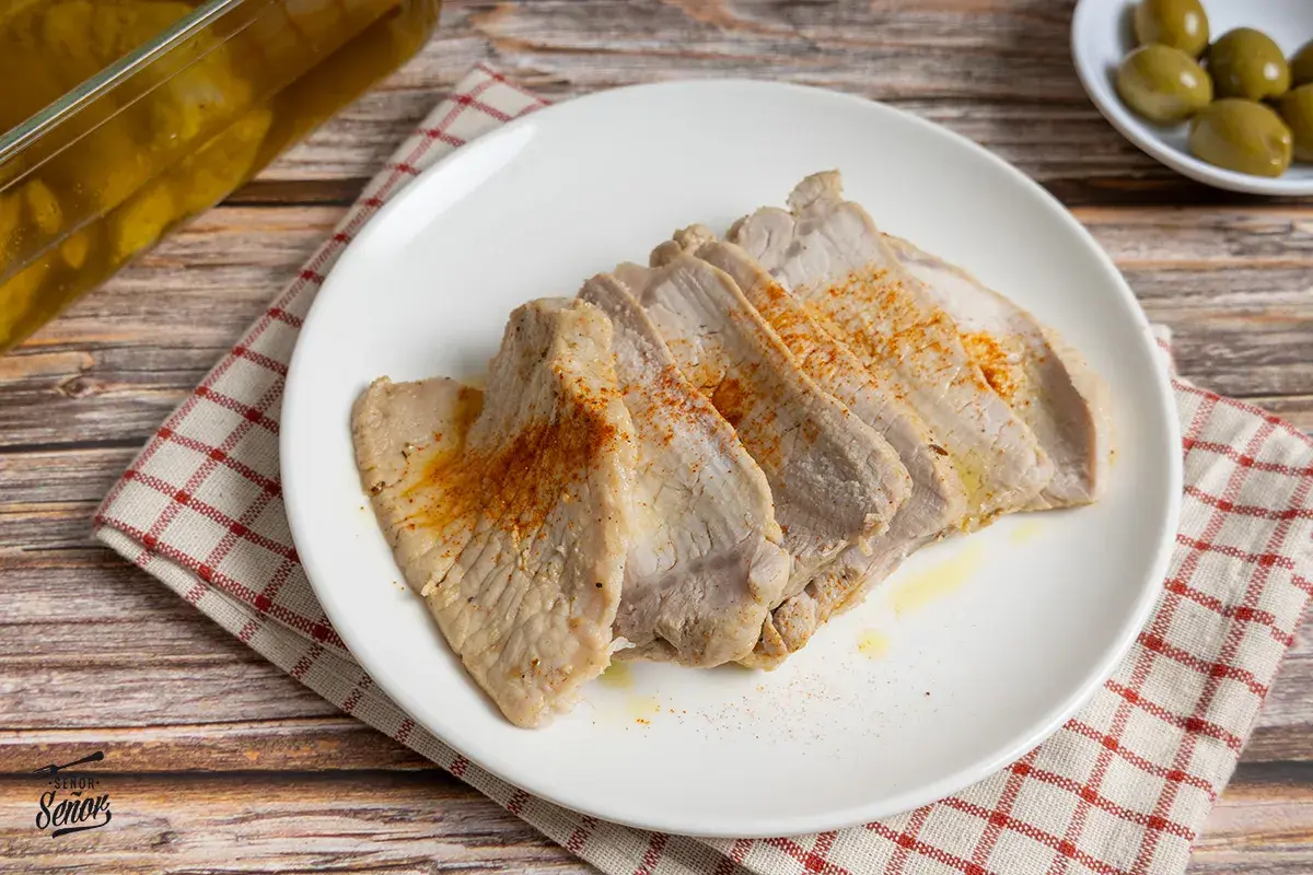 Lomo de orza tierno y jugoso, cortado en lonchas y espolvoreado con pimentón. Servido en plato blanco sobre mantel a cuadros.