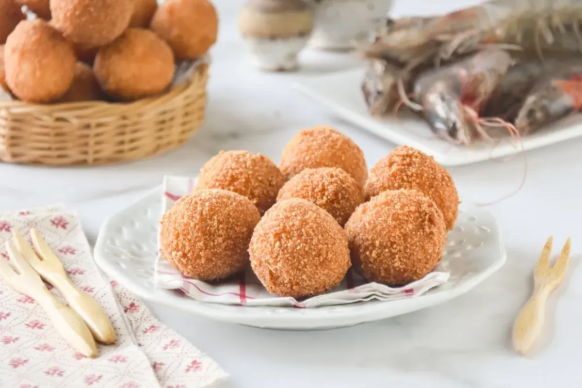 Croquetas de gambas doradas y crujientes en un plato blanco, con más en una cesta de mimbre al fondo y gambas frescas.