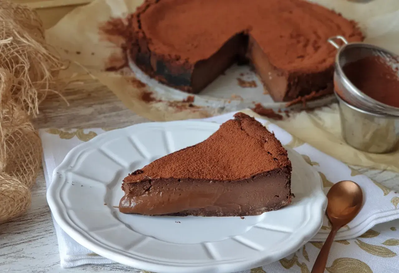 Una rebanada de tarta de chocolate, cubierta de cacao en polvo, reposa en un plato blanco. El postre parece cremoso y delicioso.