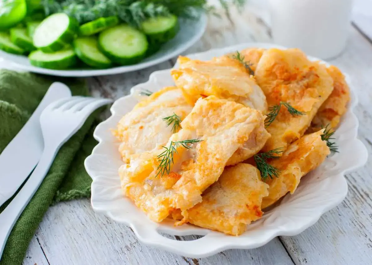 Delicioso cachopo de merluza rebozado, servido en plato blanco con eneldo fresco. Acompañado de rodajas de pepino.