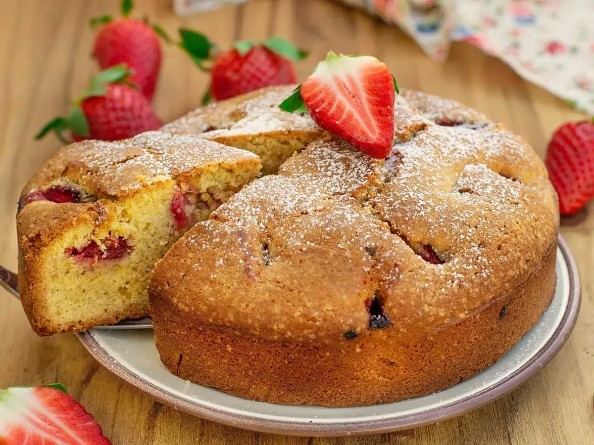 Delicioso bizcocho de fresa, espolvoreado con azúcar glas y decorado con una fresa fresca. Se ve una porción cortada, revelando trozos de fresa en su interior.