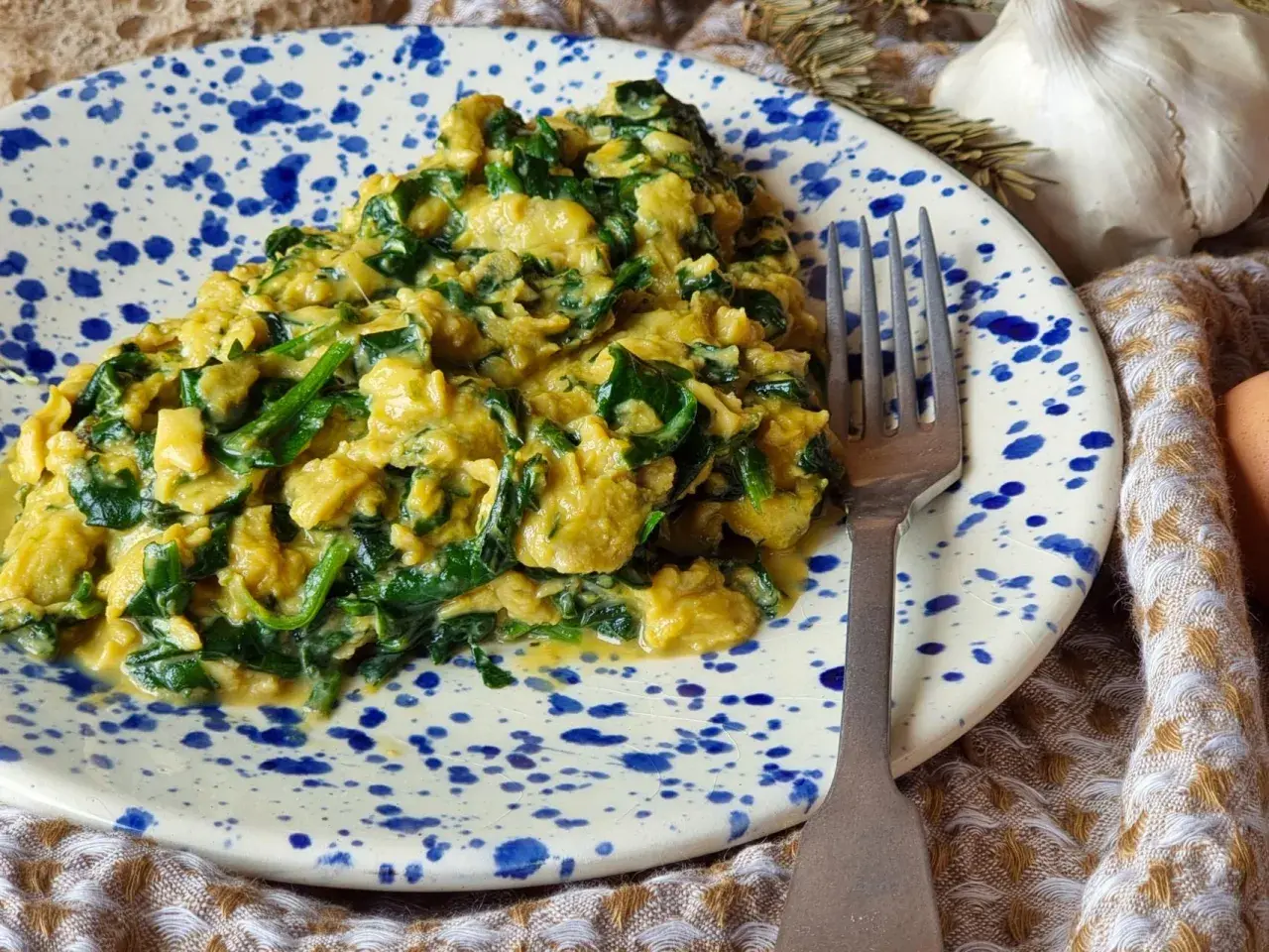 Un delicioso revuelto de espinacas cremoso, servido en un plato con salpicaduras azules. Un tenedor plateado descansa a un lado, listo para disfrutar.