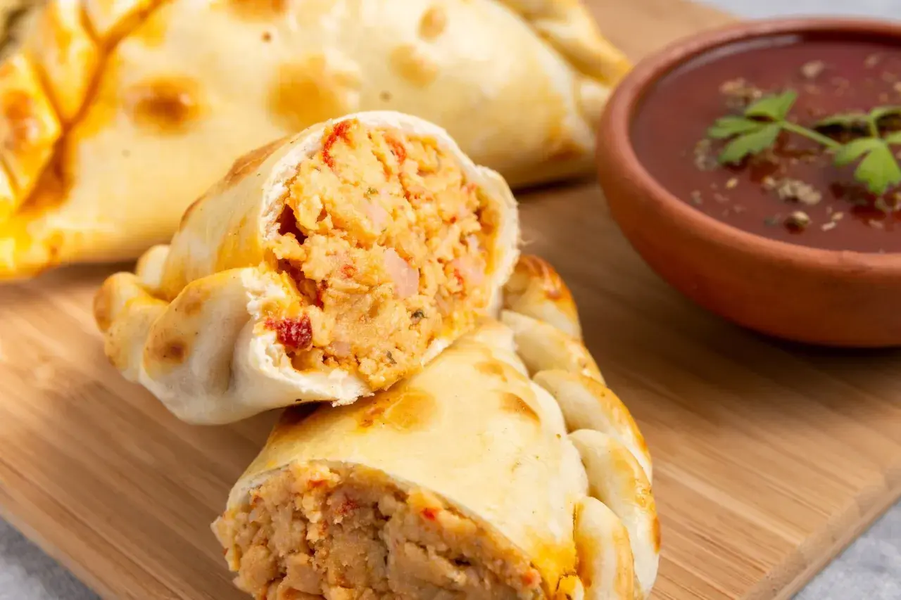 Empanada de pollo recién horneada, cortada por la mitad, revela un relleno jugoso. A su lado, un cuenco de salsa roja con perejil.
