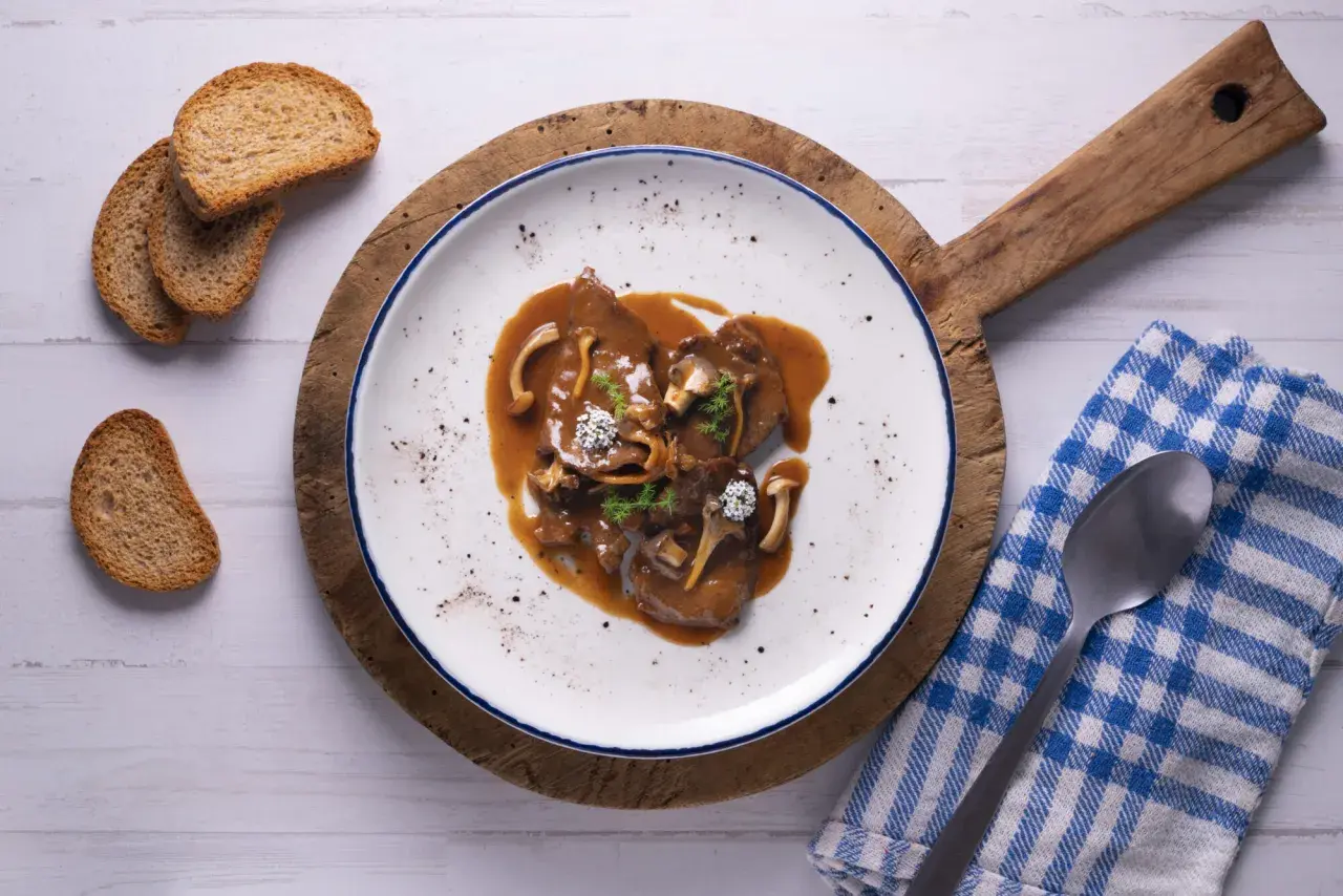Fricandó de vedella amb bolets i una salsa espessa, servit en un plat blanc amb vores blaves, acompanyat de torradetes.