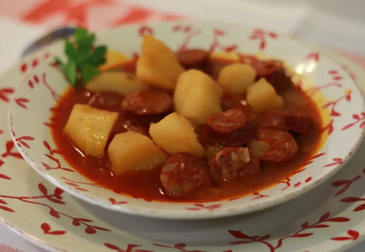 Un plato humeante de patatas a la riojana, con trozos de chorizo y patata en una salsa rojiza, decorado con una ramita de perejil.