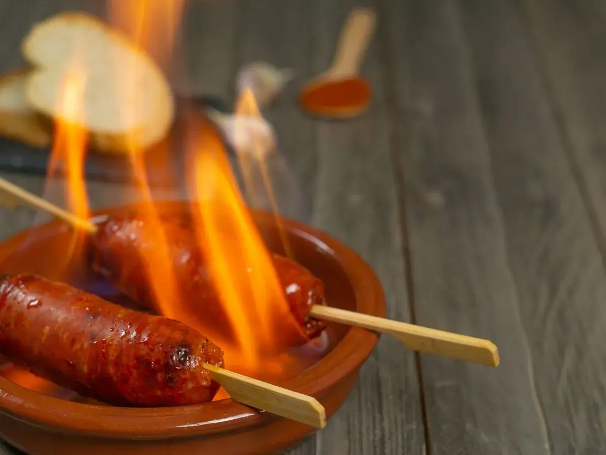 Chorizo al infierno, dos salchichas en brochetas ardiendo en una cazuela de barro, con pan y pimentón al fondo.