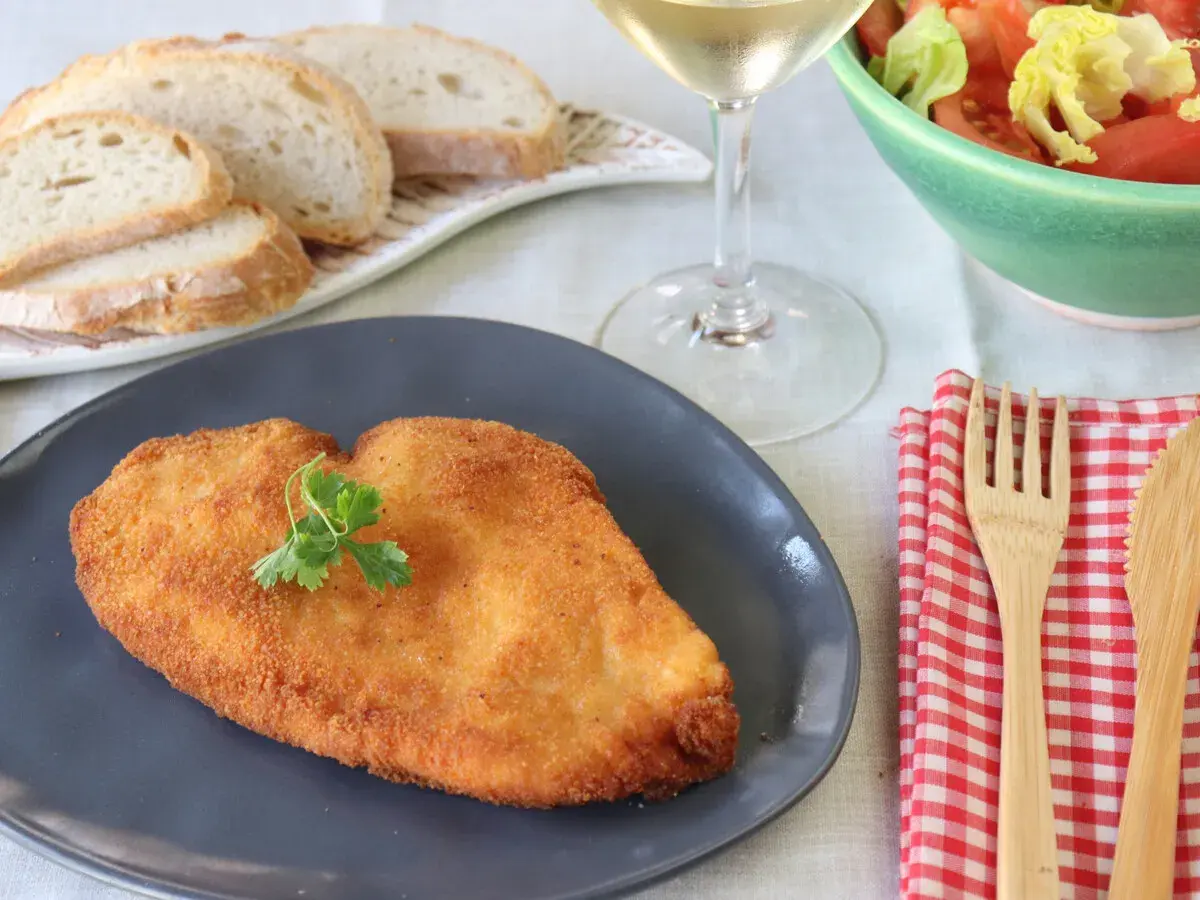 Un delicioso escalope de pollo dorado, servido con pan, ensalada y vino blanco.
