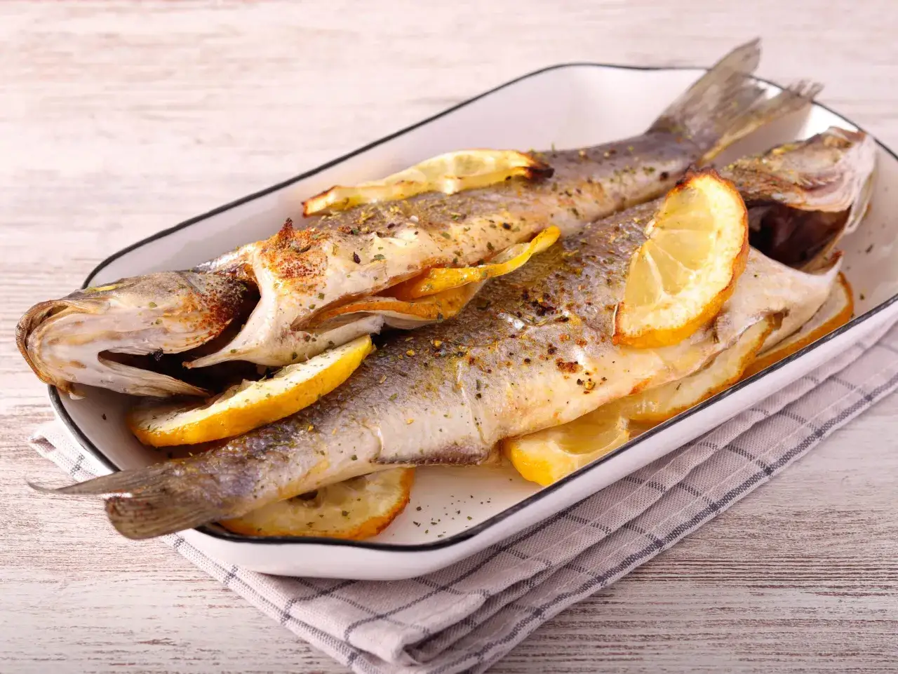 Dos lubinas al horno con patatas, doradas y jugosas, adornadas con rodajas de lim&oacute;n y hierbas arom&aacute;ticas.