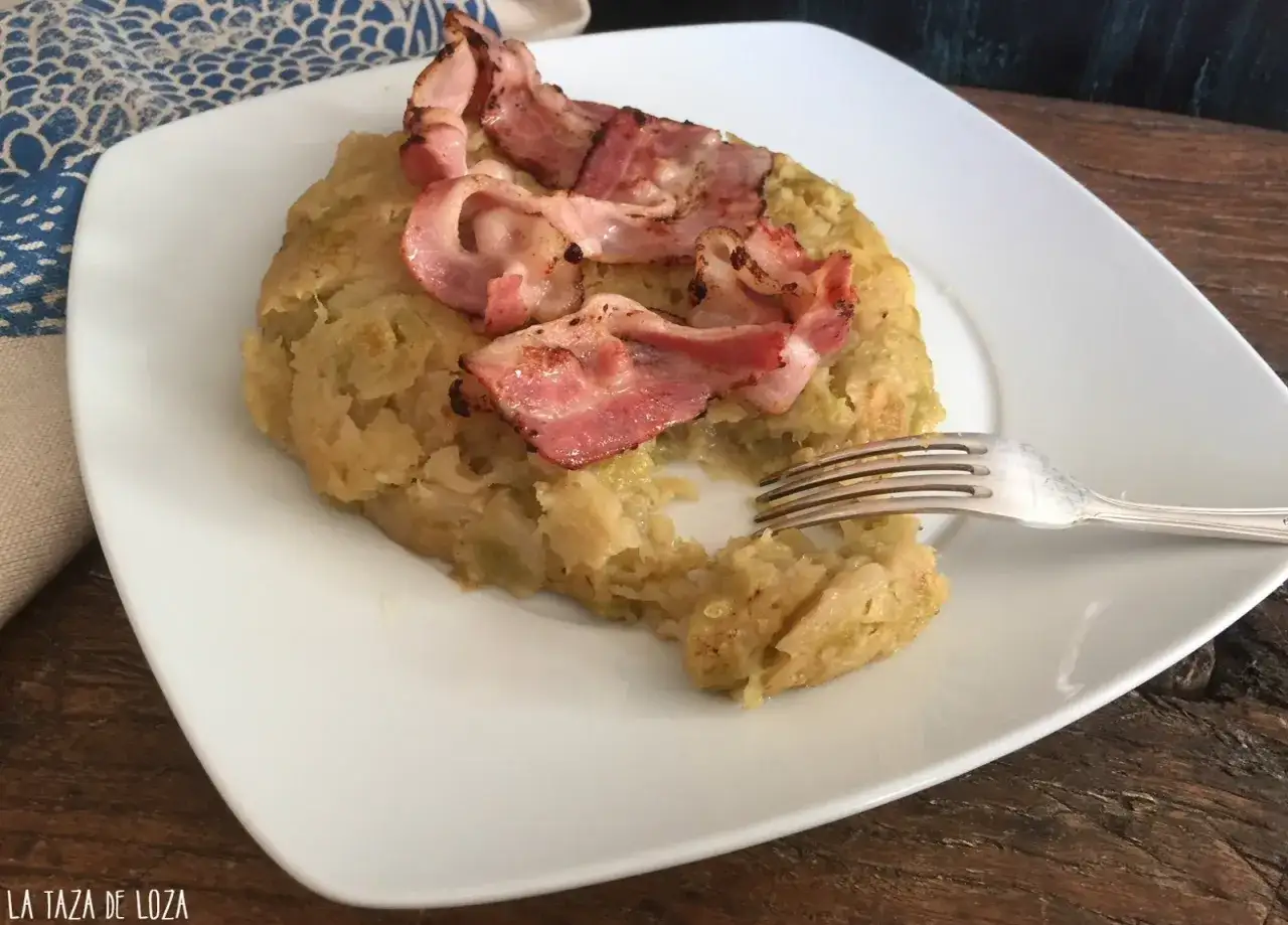 Un plato de trinxat de monta&ntilde;a, con trozos de col y patata, coronado con crujientes tiras de bacon. Un tenedor listo para probarlo.