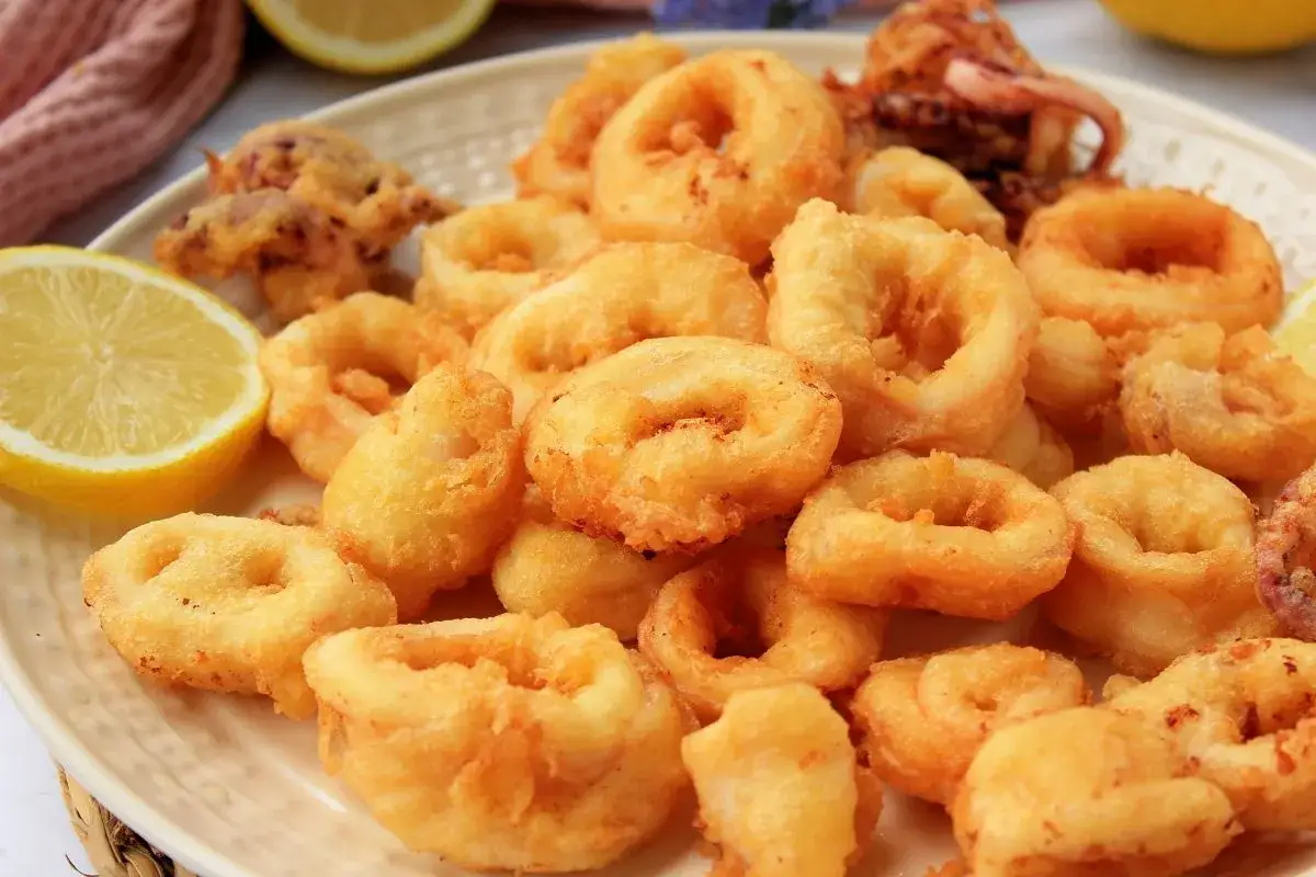 Crujientes aros de calamar a la romana dorados, servidos en un plato blanco con gajos de lim&oacute;n fresco.