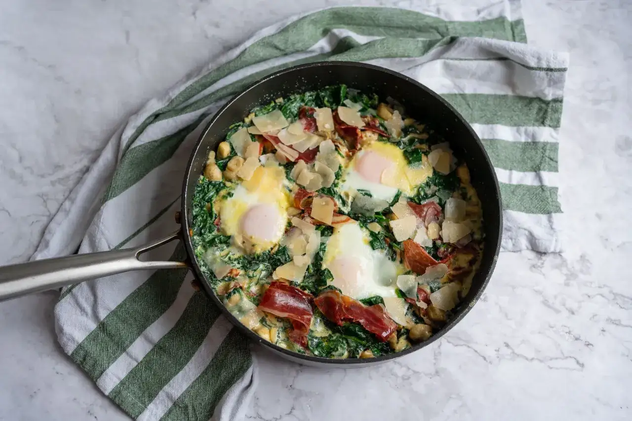 Revuelto de espinacas con huevos pochados, jam&oacute;n serrano y virutas de queso parmesano en una sart&eacute;n.