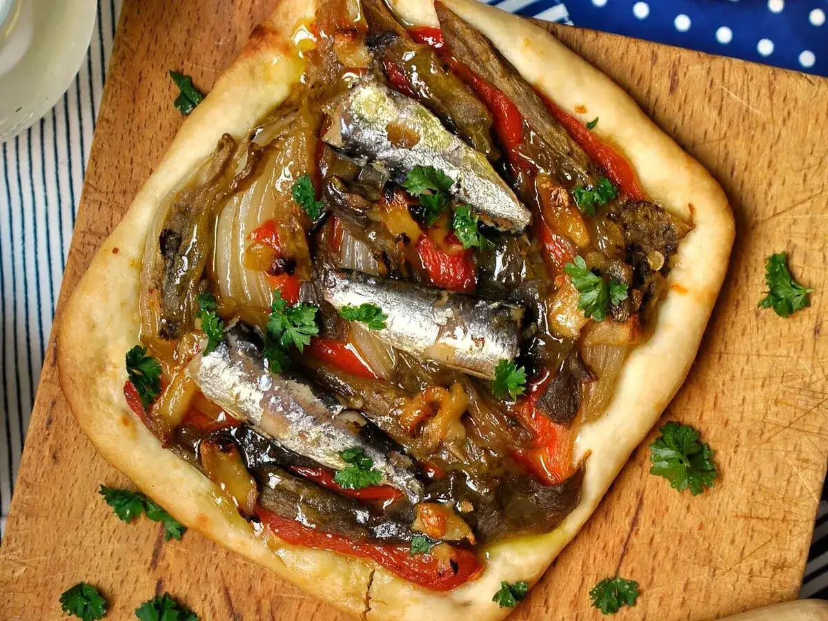 Deliciosa coca de verduras con sardinas y perejil fresco, servida sobre tabla de madera.