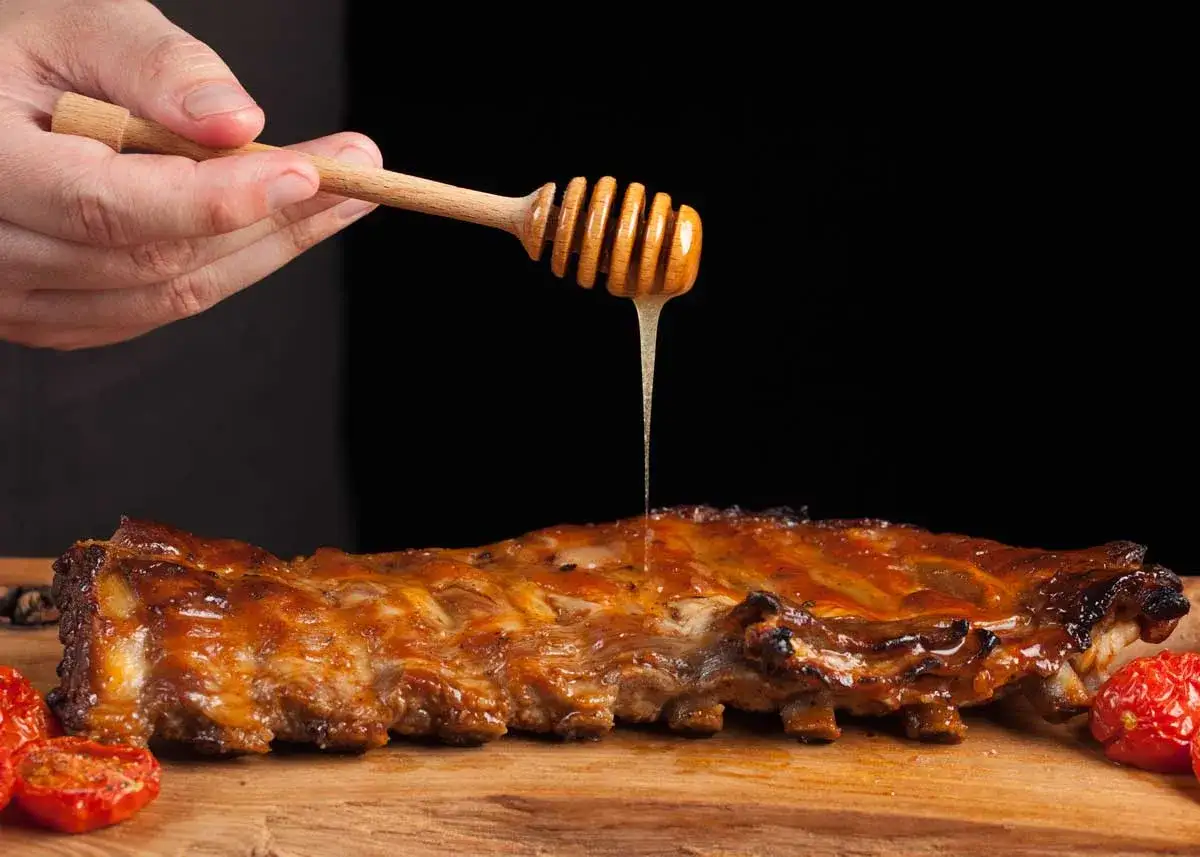 Costillas a la miel jugosas, ba&ntilde;adas con miel dorada de un palillo de madera, acompa&ntilde;adas de tomates asados.
