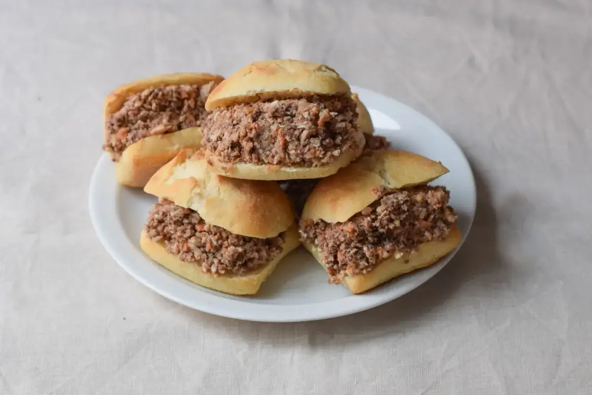 Cinco montaditos de pringa, rellenos de carne desmenuzada, apilados en un plato blanco sobre un mantel de lino.