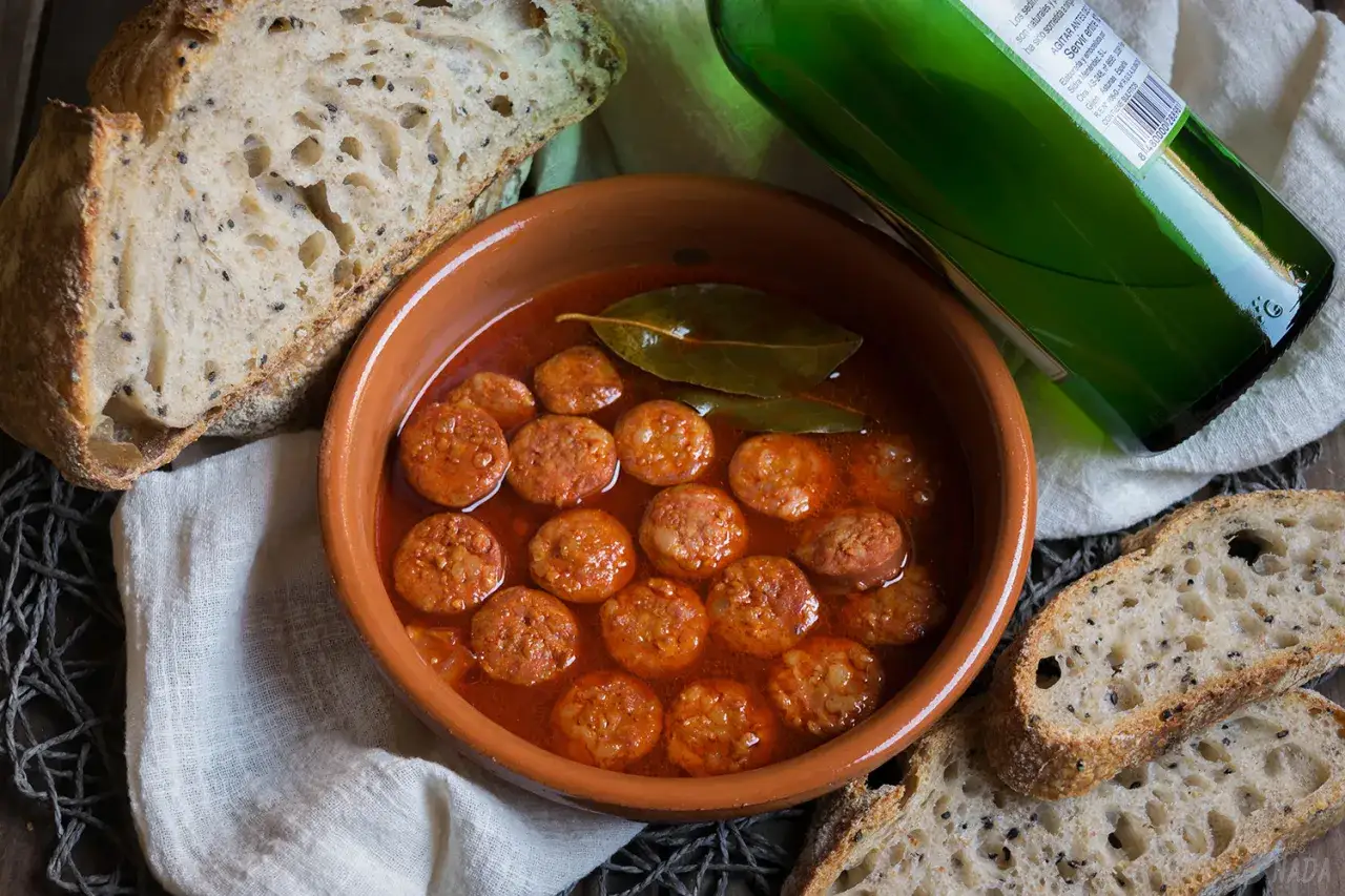 Taz&oacute;n de chorizos a la sidra, con pan r&uacute;stico y una botella de sidra verde.