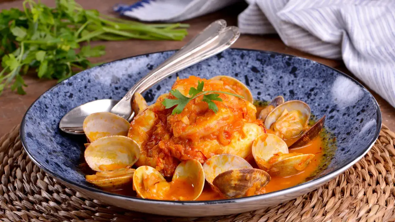 Merluza a la marinera con almejas y gambas, un plato de pescado en salsa roja con mariscos, decorado con perejil.