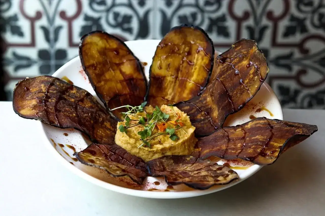 Berenjenas con miel, crujientes y caramelizadas, dispuestas alrededor de un cremoso hummus decorado con hierbas frescas.