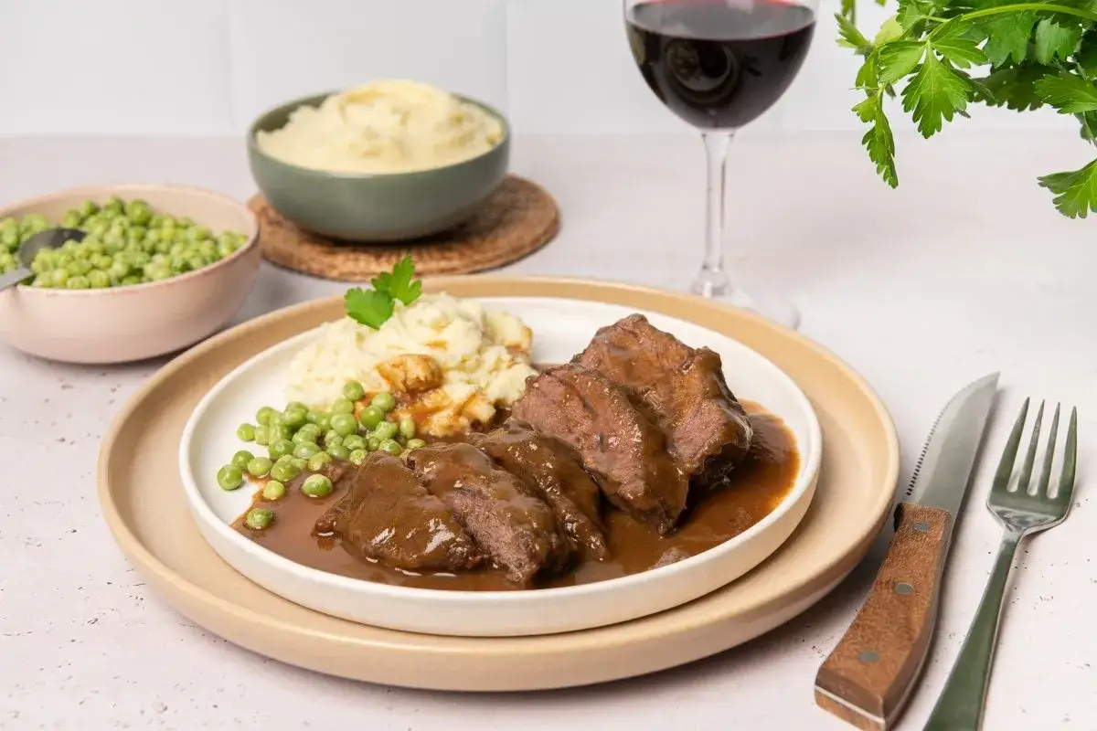 Carrilleras de ternera estofadas en salsa, servidas con pur&eacute; de patatas y guisantes. Un plato reconfortante con una copa de vino tinto.