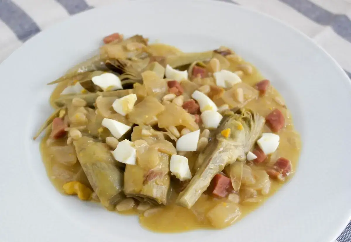 Plato de alcachofas con jam&oacute;n, huevo duro y pi&ntilde;ones, servido en un plato blanco.