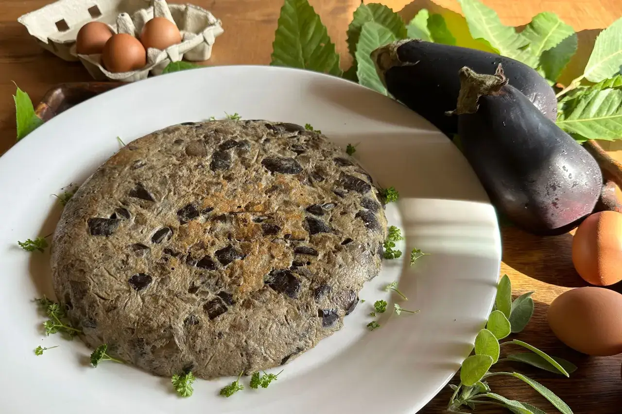 Tortilla de berenjena r&uacute;stica, con trozos visibles de la verdura, decorada con perejil fresco. Acompa&ntilde;ada de berenjenas crudas y huevos.