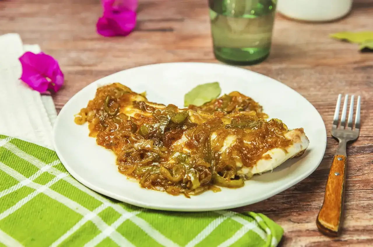 Un plato de urta a la rote&ntilde;a, con pescado blanco cubierto de cebolla caramelizada y pimientos verdes. Servido en un plato blanco sobre una mesa de madera.