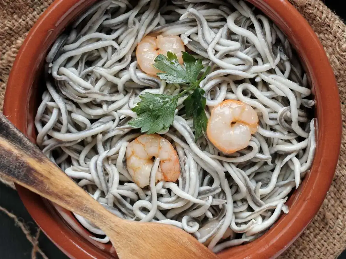 Gulas al ajillo, un plato tentador de angulas con gambas y perejil, servido en cazuela de barro con cuchara de madera.