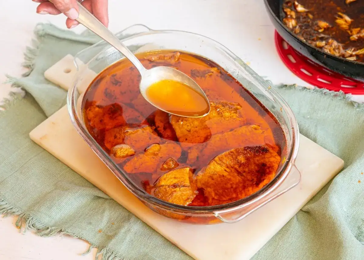 Cuchara sirviendo jugo dorado sobre trozos de lomo en manteca, cocin&aacute;ndose en un recipiente de vidrio.